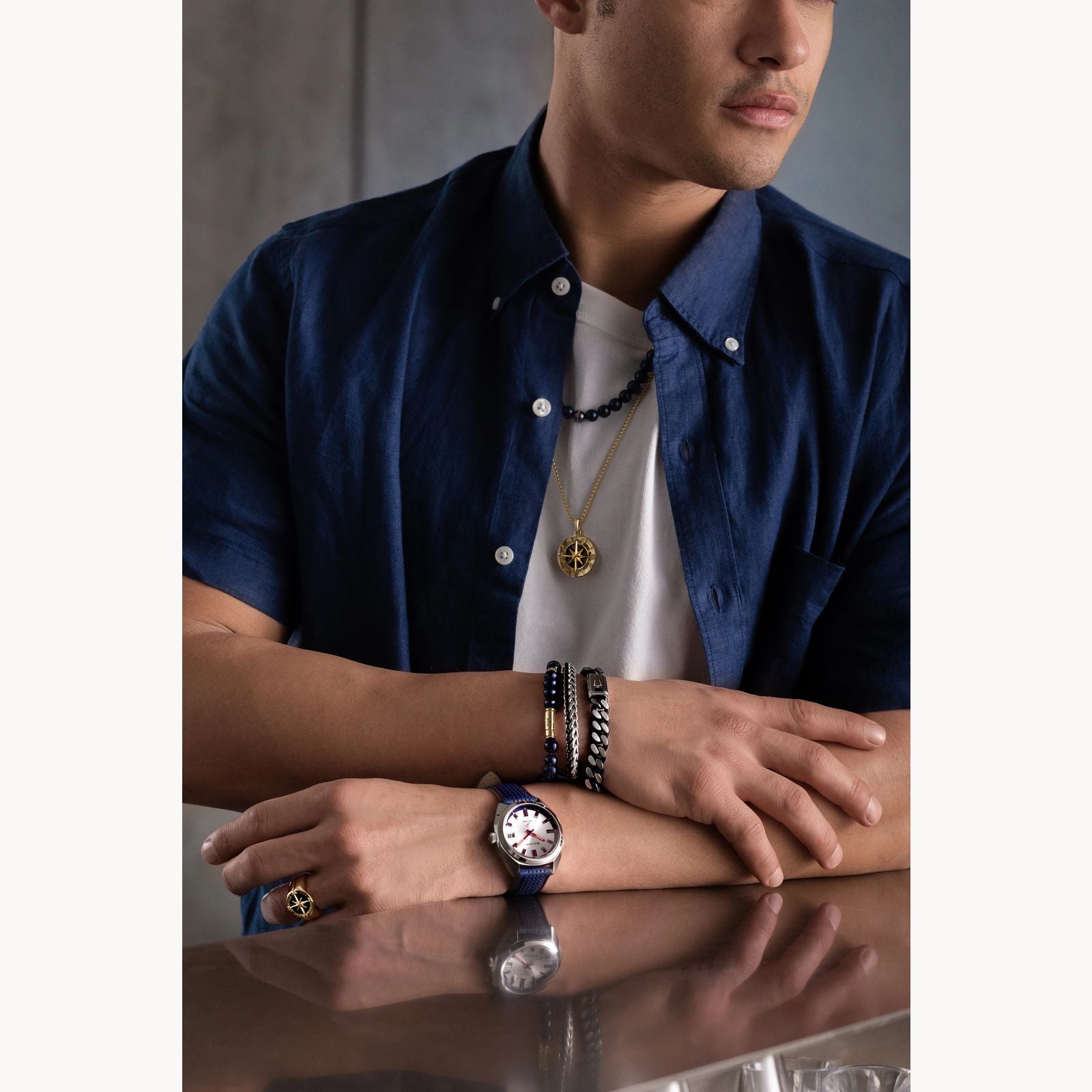 A man in a navy short-sleeve shirt wears layered necklaces, rings, a watch, and the Bulova Watch Company Curb Chain Bracelet in stainless steel, resting his arms on a reflective surface with his face partially visible and looking away.