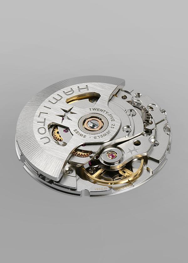 Close-up of the movement mechanism of the Hamilton Khaki Navy Scuba Automatic Watch, revealing intricate gears, metallic components, and engraved Hamilton branding on a circular silver rotor against a plain gray background. The watch’s durable design is enhanced by a ceramic bezel and 300m water resistance.