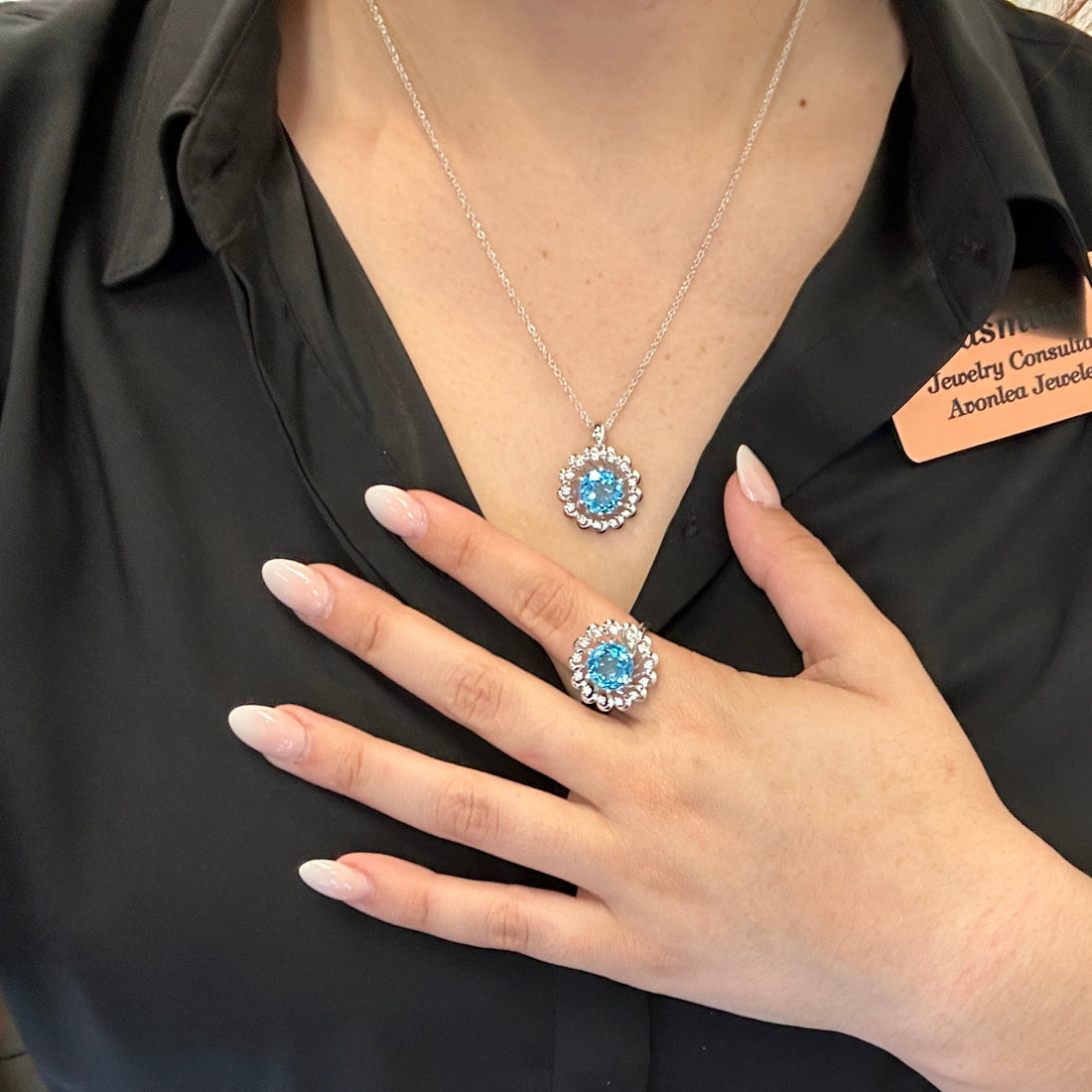 A person wearing a black blouse showcases a matching jewelry set from Allison Kaufman. The standout pieces include the "Swiss Blue Topaz and Diamonds 14K White Gold Necklace" paired with a magnificent ring, both adorned with Swiss blue topaz gemstones encircled by diamond accents. Their light pink manicured nails elegantly complete the sophisticated ensemble.