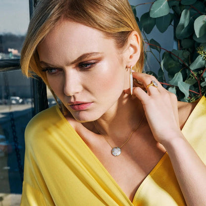 A woman stands by a window gazing downward, wearing a yellow blouse and adorned with Freida Rothman's Petals and Pavé Small Pendant Necklace, hoop earrings, and a ring. Her blonde hair glows softly in the light against the Brooklyn skyline backdrop with leaves trailing behind her.