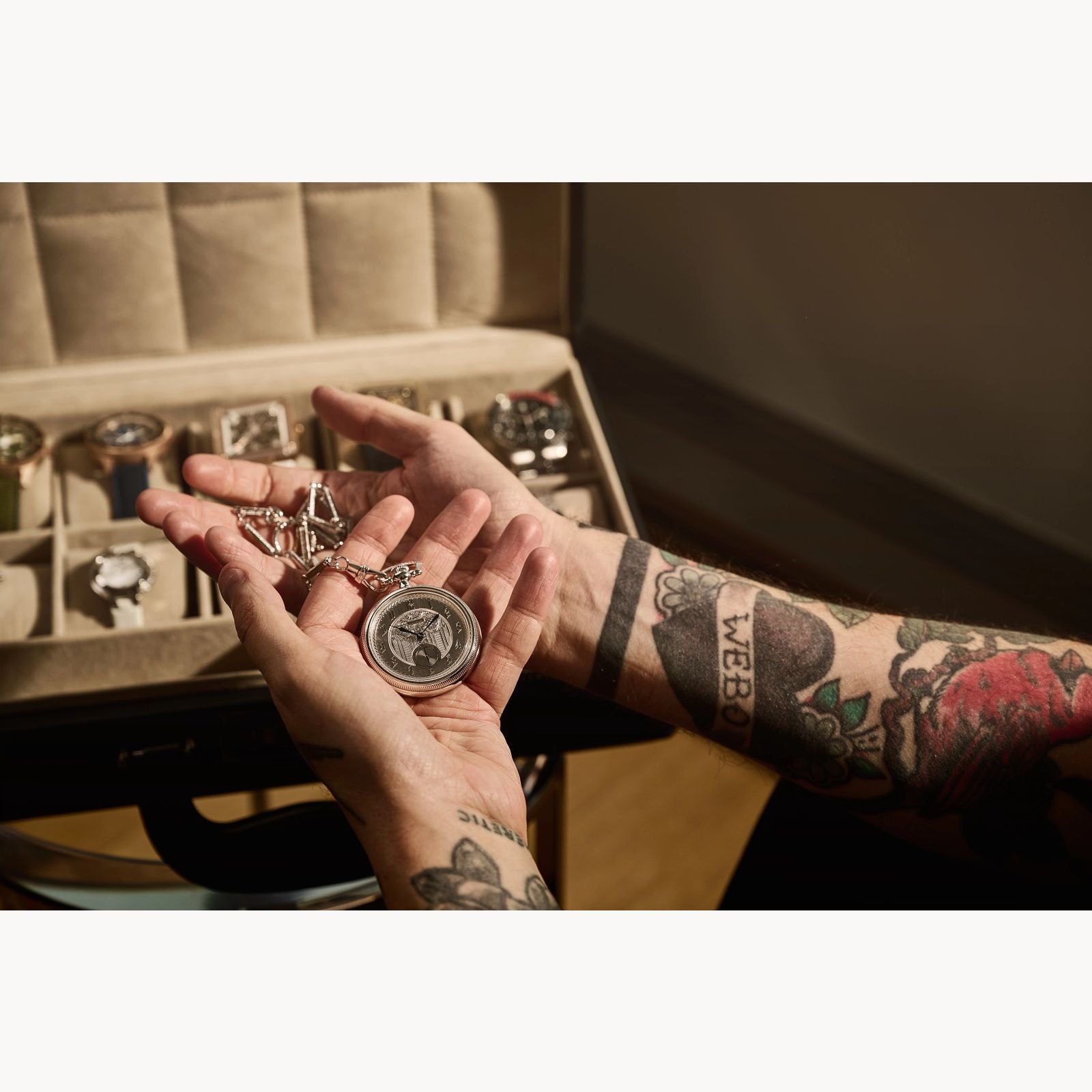 A tattooed person holds an open Bulova Limited Edition Silver-Tone Stainless Steel Chain Pocket Watch by Bulova Watch Company above a jewelry box of wristwatches, showcasing the sophistication of this exclusive timepiece.