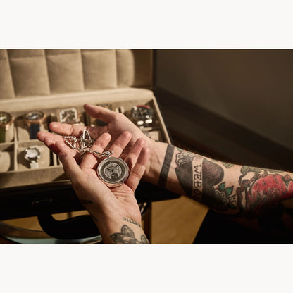 A tattooed person holds an open Bulova Limited Edition Silver-Tone Stainless Steel Chain Pocket Watch by Bulova Watch Company above a jewelry box of wristwatches, showcasing the sophistication of this exclusive timepiece.