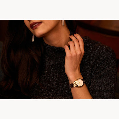 A woman wearing a dark, sparkly top and drop earrings touches her neck, showcasing the Bulova Watch Company Ladies Surveyor in stainless steel with a sapphire crystal and MOP dial. Her maroon lipstick and dark hair are partially visible.