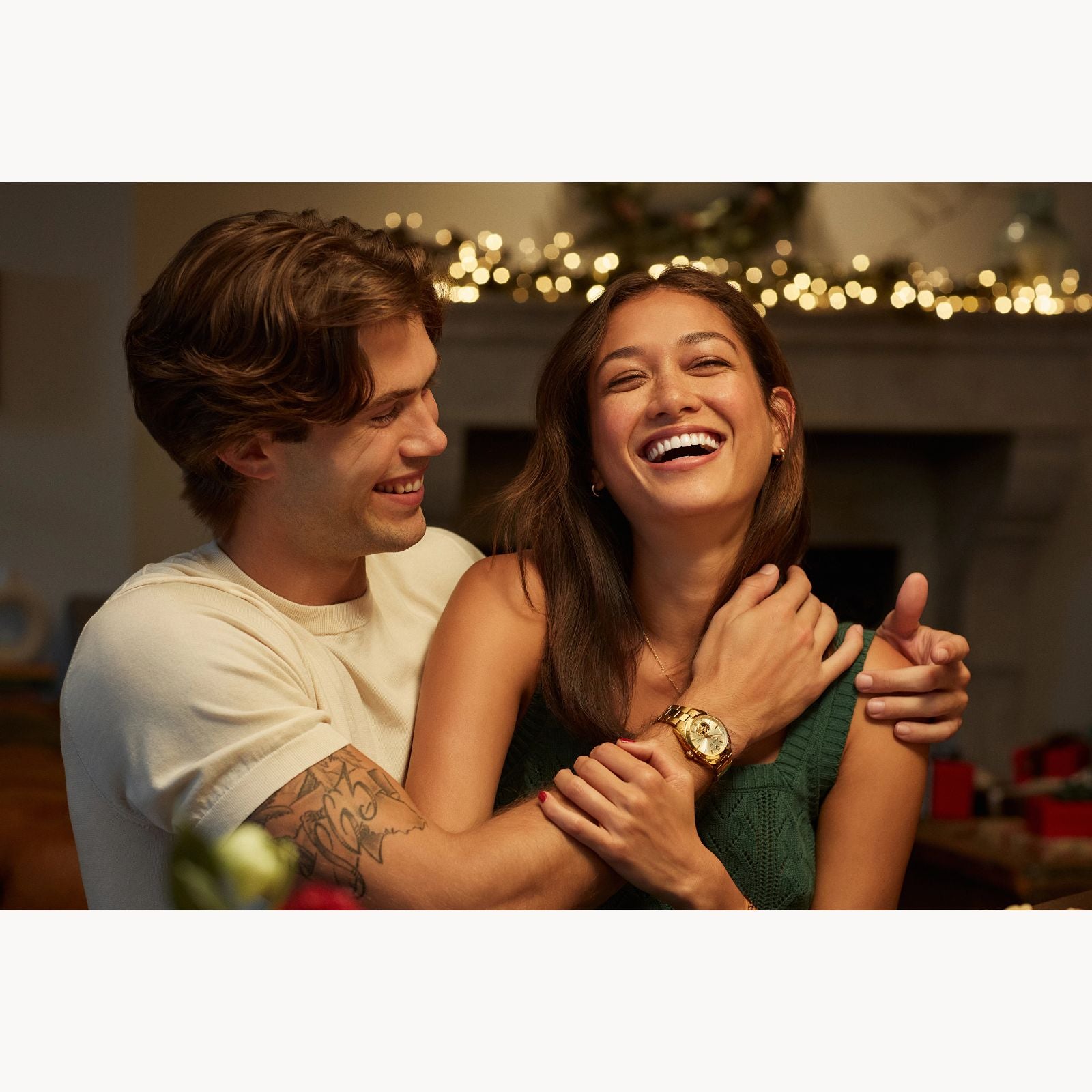 A smiling couple embraces and laughs indoors under festive string lights. The man proudly wears the Bulova Watch Company Surveyor M Y BR CH AUTO, its sleek, automatic design adding sophistication to the joyful moment.