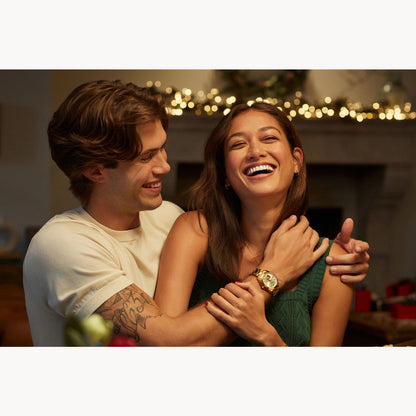 A smiling couple embraces and laughs indoors under festive string lights. The man proudly wears the Bulova Watch Company Surveyor M Y BR CH AUTO, its sleek, automatic design adding sophistication to the joyful moment.
