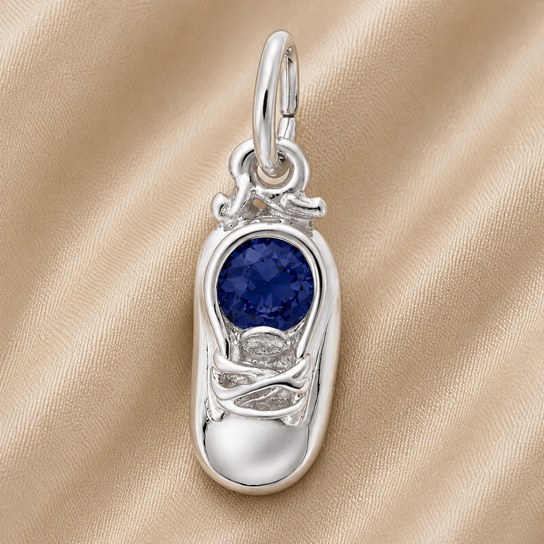 The Rembrandt Charms Baby Shoe Birthstone (September) pendant, crafted in sterling silver and adorned with a round, deep blue gemstone at its center, makes a perfect September birthstone charm or meaningful birthday gift. Shown resting on soft beige satin fabric, this intricately detailed baby shoe features delicate laces and a small loop for easy attachment to your favorite chain.