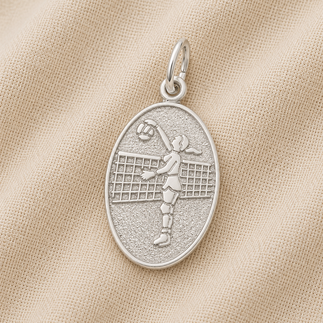 The Rembrandt Charms Volleyball Player Oval Disc Charm in sterling silver showcases an engraved image of a volleyball player jumping to hit the ball over a net, displayed on a textured beige fabric background—an elegant example of sterling silver volleyball jewelry and the perfect charm for any enthusiast.