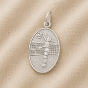 The Rembrandt Charms Volleyball Player Oval Disc Charm in sterling silver showcases an engraved image of a volleyball player jumping to hit the ball over a net, displayed on a textured beige fabric background—an elegant example of sterling silver volleyball jewelry and the perfect charm for any enthusiast.