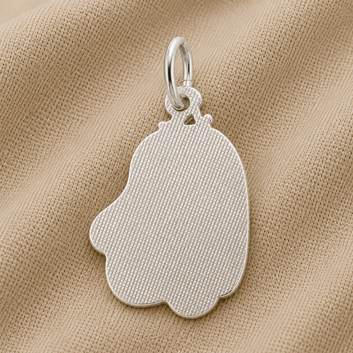 The “Cute Ghost Charm- Sterling Silver” by Rembrandt Charms, featuring a textured finish and an abstract bell-like silhouette that evokes a playful ghost, is elegantly displayed on beige fabric—a sophisticated take on Halloween jewelry.