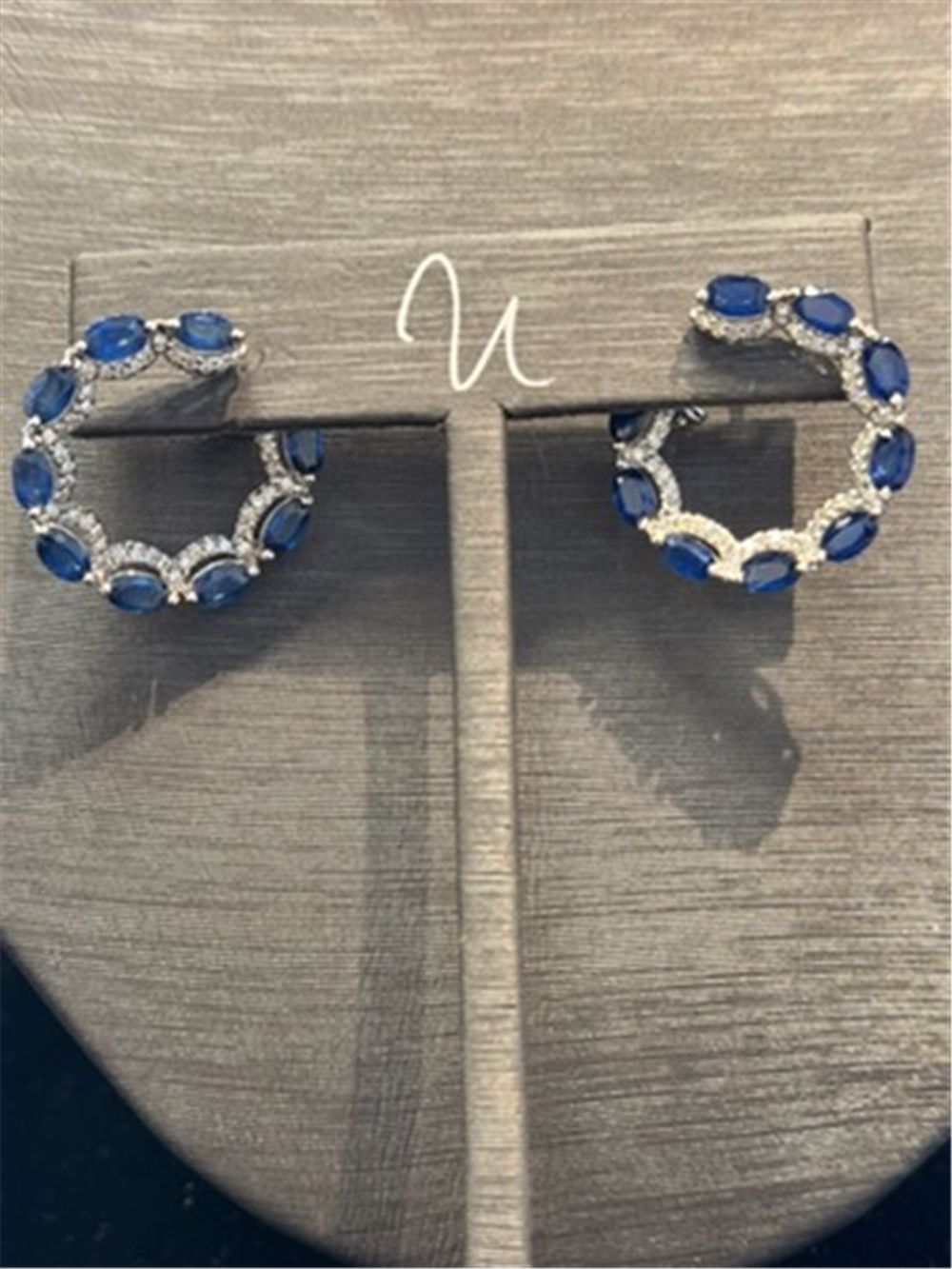 The Shappire and Diamond Wrap Around Hoops by Ryan Gems feature alternating blue sapphires and diamonds in 14k white gold, displayed on a gray jewelry stand.