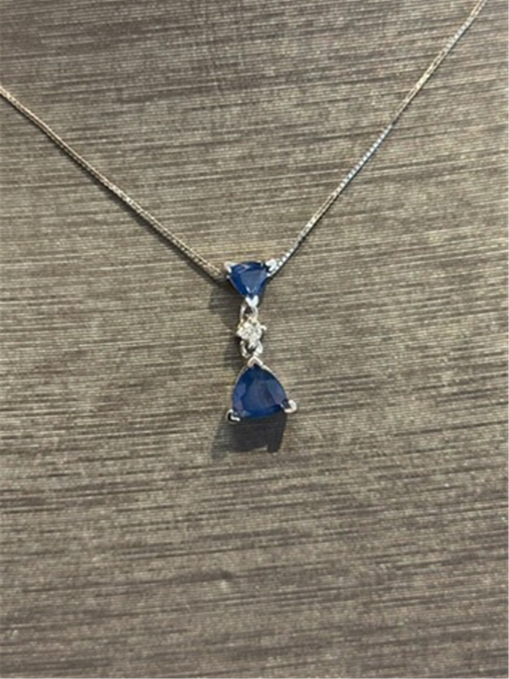 The Ryan Gems Trillion Cut Sapphire Drop Pendant in 14k white gold features two trillion-cut blue sapphires and a small central clear stone, shown on a textured gray surface.