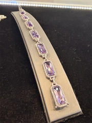 The Ryan Gems Diamond and Amethyst Large Tennis Bracelet in 14k white gold features seven rectangular purple gemstones with diamond accents, shown on a gray stand against a black background.