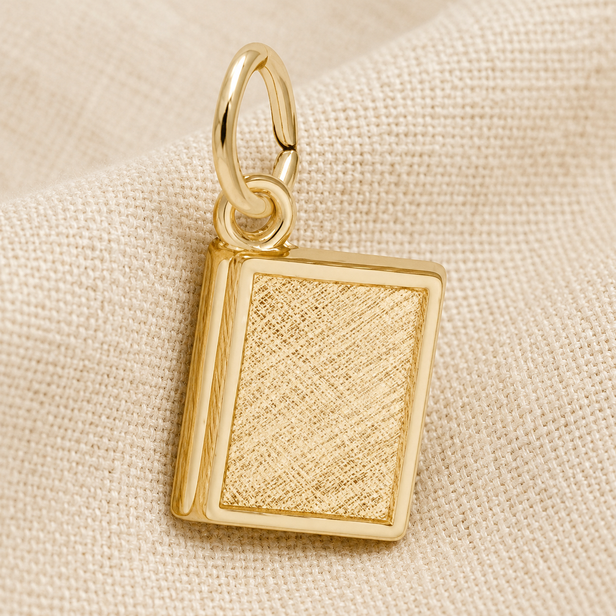 A small, rectangular gold plated charm from Rembrandt Charms, designed to resemble a textured book, rests on a beige fabric background—the perfect Book Charm for literary enthusiasts seeking personalized jewelry.