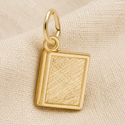 A small, rectangular gold plated charm from Rembrandt Charms, designed to resemble a textured book, rests on a beige fabric background—the perfect Book Charm for literary enthusiasts seeking personalized jewelry.