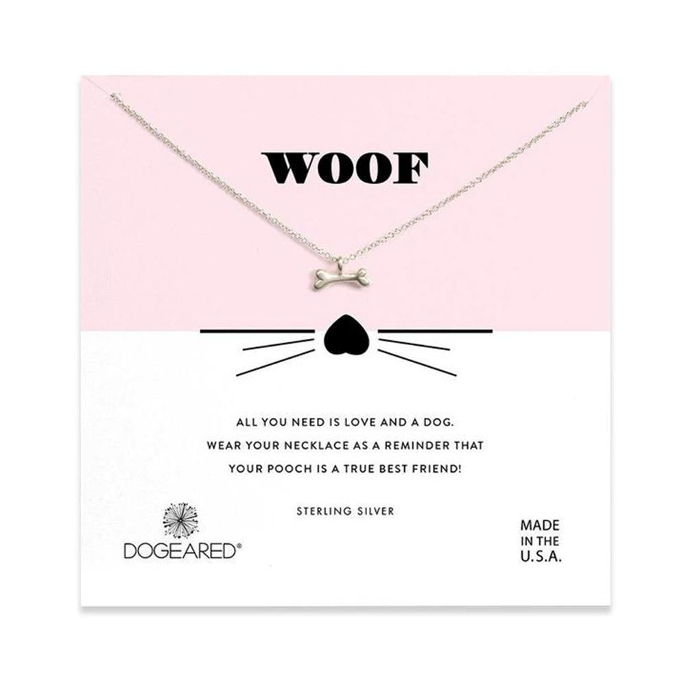 The Woof /SS pendant necklace by Dogeared features a silver bone-shaped charm and is presented on a card with a pink and white background. The card reads "WOOF" at the top, accompanied by a small heart with whiskers illustration. Below, it states, "All you need is love and a dog. Wear your necklace as a reminder that your pooch is a true best friend!" Crafted from sterling silver in the U.S.A.