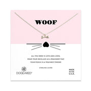 The Woof /SS pendant necklace by Dogeared features a silver bone-shaped charm and is presented on a card with a pink and white background. The card reads "WOOF" at the top, accompanied by a small heart with whiskers illustration. Below, it states, "All you need is love and a dog. Wear your necklace as a reminder that your pooch is a true best friend!" Crafted from sterling silver in the U.S.A.