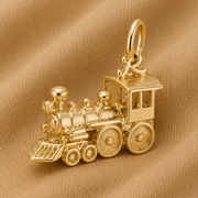 The Rembrandt Charms Train Engine Charm – Gold Plated is displayed on a soft, tan fabric background, making it an ideal piece of locomotive jewelry for collectors and train enthusiasts.