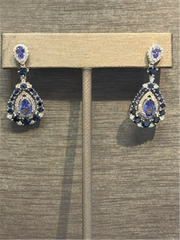 The Ryan Gems Tanz, Saph, and Dia Dangles are 14k white gold teardrop earrings featuring blue sapphires and diamonds, shown on a beige stand against a textured gray background.
