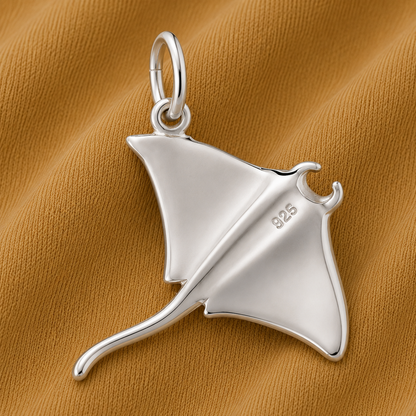 A Rembrandt Charms Manta Ray Charm in sterling silver (“925”) rests on a textured, mustard-yellow fabric background, showcasing the elegance of ocean-inspired jewelry.