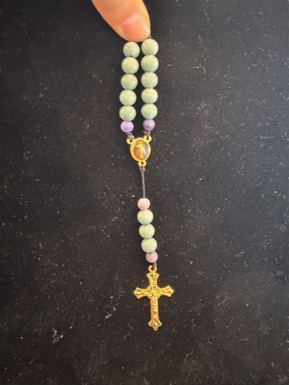 A hand holds Redemption Jewelry’s SR006 Small Jade & Frosted Dragon Agate Rosary, featuring green and purple beads, a small medallion, and gold crucifix against a dark background.