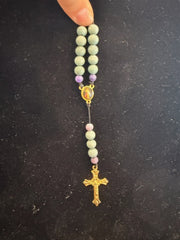 A hand holds Redemption Jewelry’s SR006 Small Jade & Frosted Dragon Agate Rosary, featuring green and purple beads, a small medallion, and gold crucifix against a dark background.