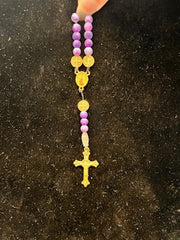 A hand holds the SR004 Small Frosted Dragon Agate Rosary by Redemption Jewelry, featuring purple frosted dragon agate beads, gold medals, and a gold crucifix against a dark textured background.