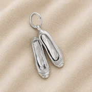 The Ballet Shoes -Sterling Silver charm by Rembrandt Charms features two elegantly crafted shoes displayed on a textured beige fabric background, making it an ideal dance keepsake for any fan of ballet jewelry.