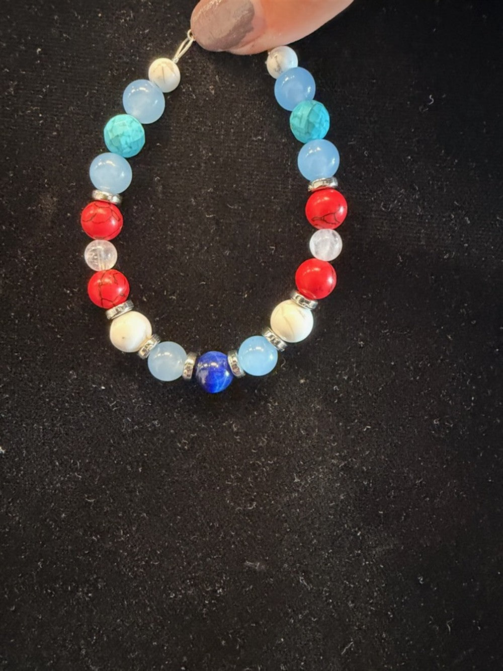 A hand with light-colored nail polish holds Redemption Jewelry’s B008 bracelet, featuring Howlite, Blue Chalcedony, Red Jasper, Rose Quartz, and Blue Tiger Eye beads separated by silver spacers against a black background.