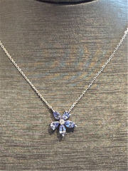 The Ryan Gems Multi Shaped Tanzanite Flower Pendant features five purple gemstone petals and a clear center on a 14k white gold necklace, displayed on a textured brown surface.