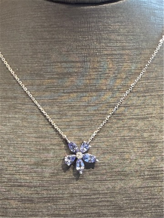 The Ryan Gems Multi Shaped Tanzanite Flower Pendant features five purple gemstone petals and a clear center on a 14k white gold necklace, displayed on a textured brown surface.