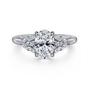A close-up of the 14K Gold Oval Diamond Engagement Ring by Gabriel & Co features a central oval diamond, flanked by six marquise and round diamonds. The sleek white gold band enhances its brilliance.