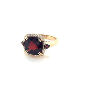 The Large Garnet and Diamonds Ring by Bellarri is a stunning 14K yellow gold piece, featuring a large, square-cut red garnet as the centerpiece. This exquisite garnet is bordered by small, clear diamonds and flanked by two smaller red stones on either side. The band completes the design with its plain gold elegance.
