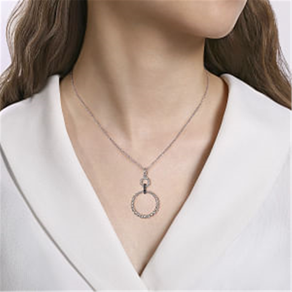 Person wearing a white V-neck top, showcasing the Gabriel & Co Sterling Silver Black Spinel Bujukan Link Drop Necklace with a round pendant hanging from a small, intertwined loop on the chain. The background is plain and light-colored, highlighting the necklace's intricate design.