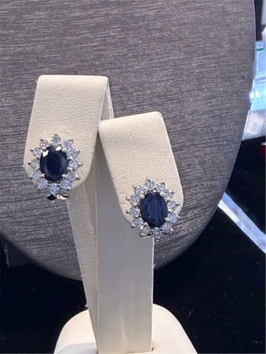 A pair of Ryan Gems Oval Sapphire Earrings w/ Halo of Diamonds set in 14k white gold, featuring dark blue sapphires and diamond accents, displayed on a cream stand against a dark background.
