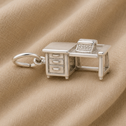 The Typewriter Desk Charm- Sterling Silver by Rembrandt Charms is crafted in the shape of an office desk with drawers and a typewriter on top, beautifully displayed on a beige fabric background—an ideal literary gift or writer jewelry piece for any wordsmith.