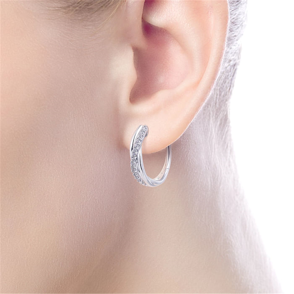 Close-up image of an ear wearing a small, elegant 925 Sterling Silver Twisted 15mm White Sapphire Huggie Earring from Gabriel & Co. The background is white, emphasizing the ear and the earring's intricate details.