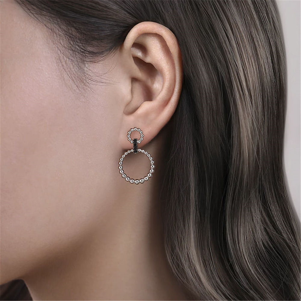 Close-up of a person wearing a pair of Gabriel & Co Sterling Silver Black Spinel Bujukan Link Drop Earrings. The elegant earrings feature two interlocking circles, each studded with small sparkling black spinel stones. The larger circle hangs below the smaller one, which is attached to the earlobe. The person's hair is long and dark.