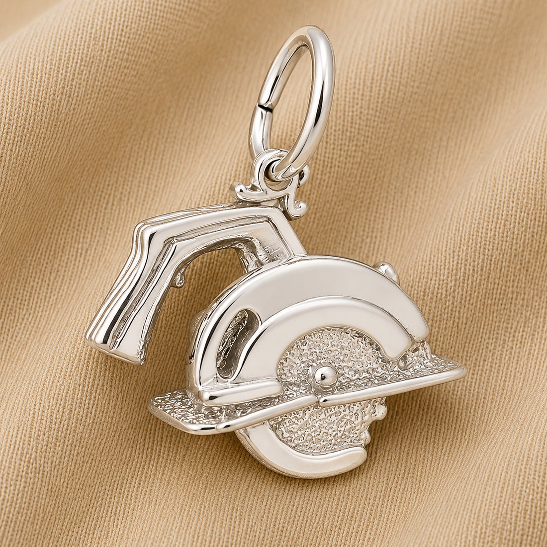 The Rembrandt Charms Electric Saw Charm, crafted in sterling silver and designed to resemble a circular saw, is displayed on light brown fabric. Featuring detailed textures like a round blade and handle, this pendant is the perfect piece of power tool jewelry and makes an ideal gift for carpenters.