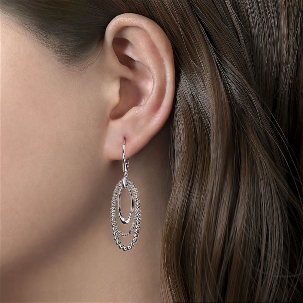 Close-up of a woman's ear adorned with an elegant silver earring from the Bujukan Collection by Gabriel & Co. The 925 Sterling Silver Bujukan and Rope Drop Earrings feature an oval hoop design with an additional smaller hoop threaded through it, connected by a row of small, round beads, adding a delicate touch to the jewelry piece.
