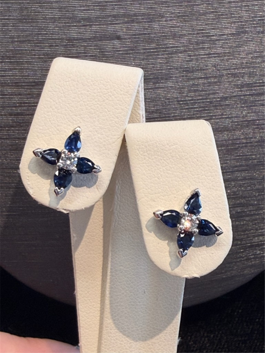 The Ryan Gems Blue Sapphire Flower Studs feature a floral design with four dark blue sapphires and a central clear stone, set in 14k white gold, displayed on a cream-colored stand.