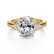 The Gabriel & Co 14K Yellow Gold Oval Lotus Diamond Engagement Ring features a 10.5x7 mm clear oval lotus diamond center stone, accented by two smaller heart-shaped clear stones on each side.