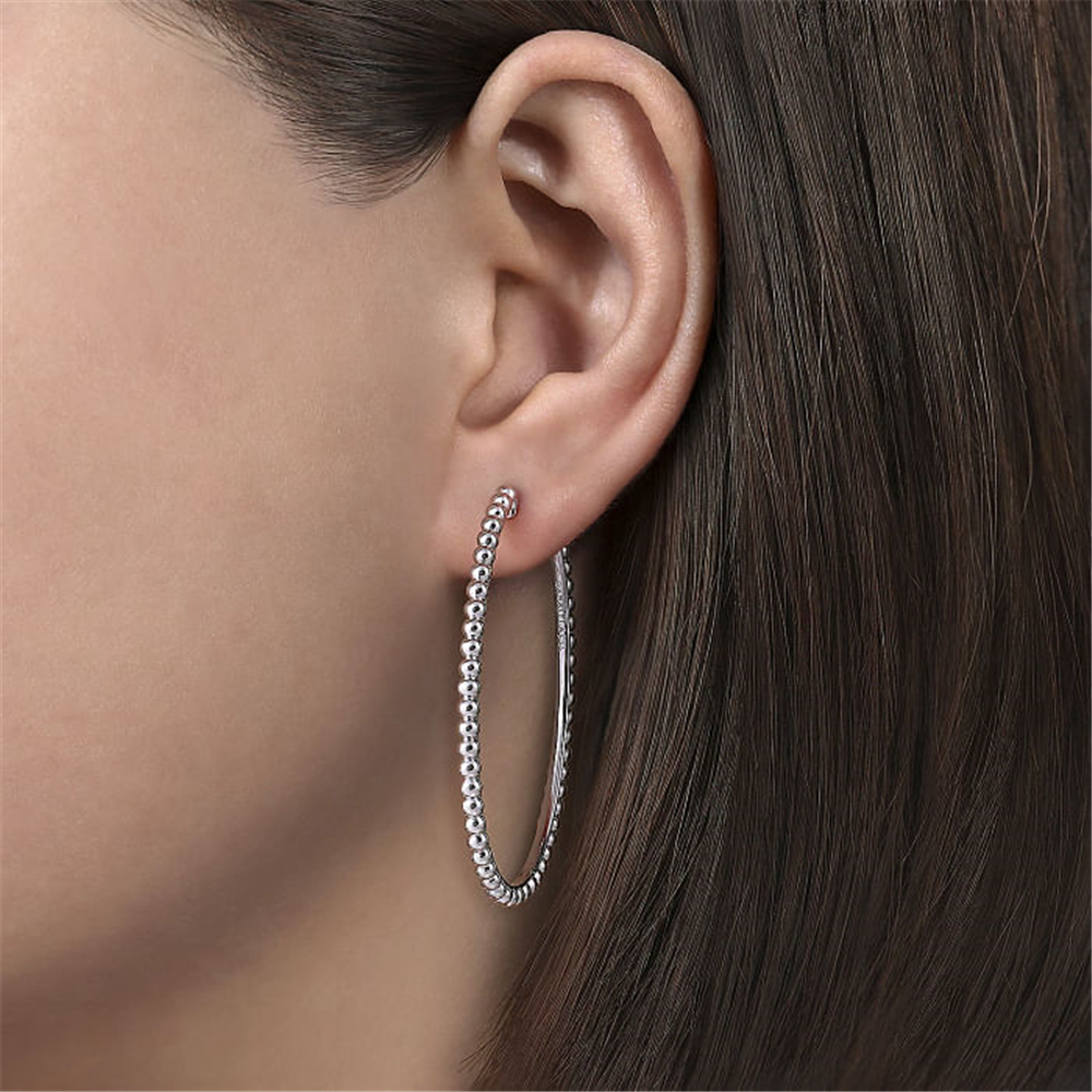 Close-up of a person wearing a large, Gabriel & Co 925 Sterling Silver Bujukan Oval Classic Hoop Earring from the Bujukan Collection. The earring features a beaded texture and is visible against the person's dark brown hair, which is straight and falls neatly behind the ear.