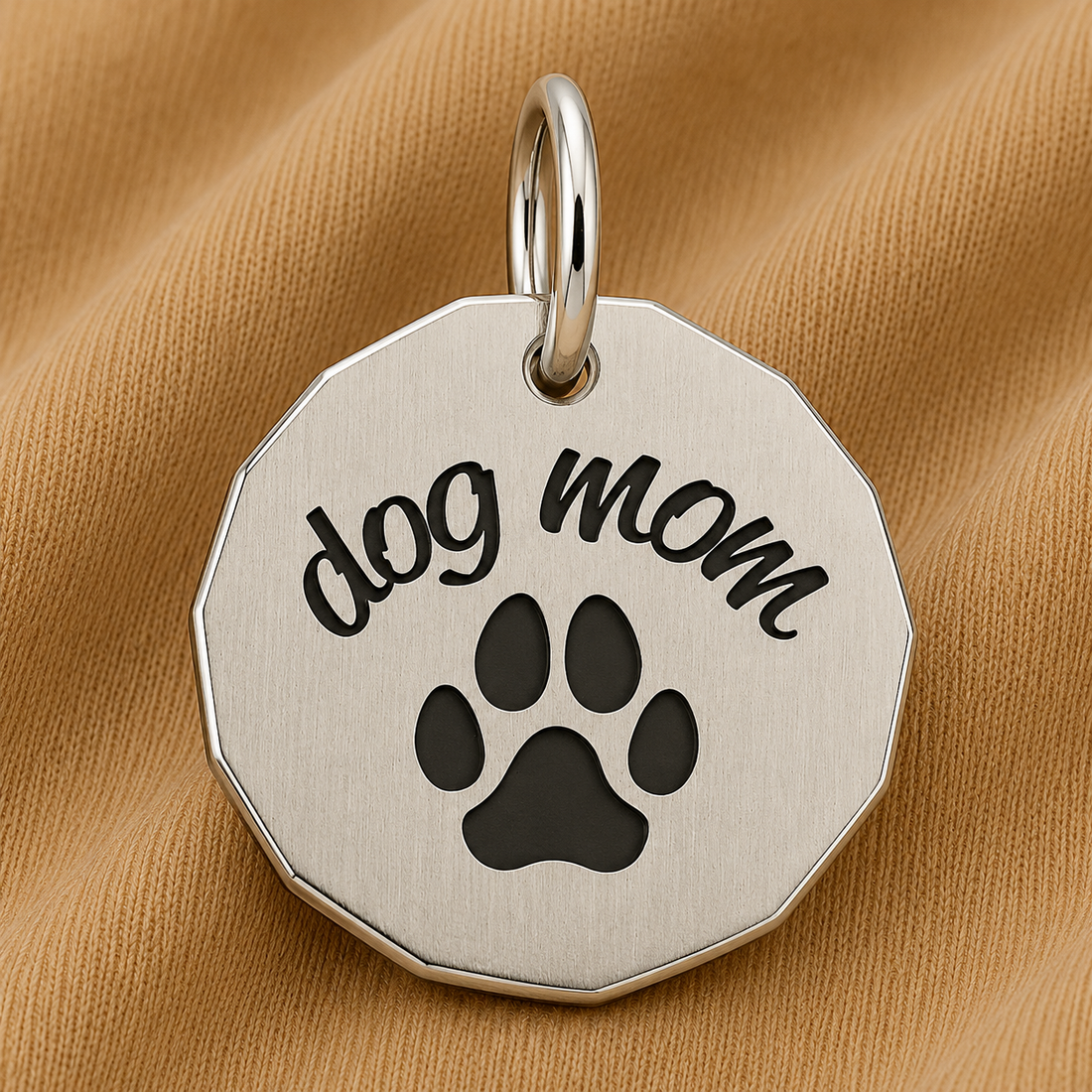 The Dog Mom Charm in sterling silver by Rembrandt Charms features “dog mom” engraved above a black paw print, displayed on light brown fabric—making it an ideal gift for pet parents and the perfect piece of paw print jewelry for any dog-loving mom.