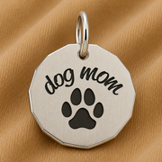 The Dog Mom Charm in sterling silver by Rembrandt Charms features “dog mom” engraved above a black paw print, displayed on light brown fabric—making it an ideal gift for pet parents and the perfect piece of paw print jewelry for any dog-loving mom.