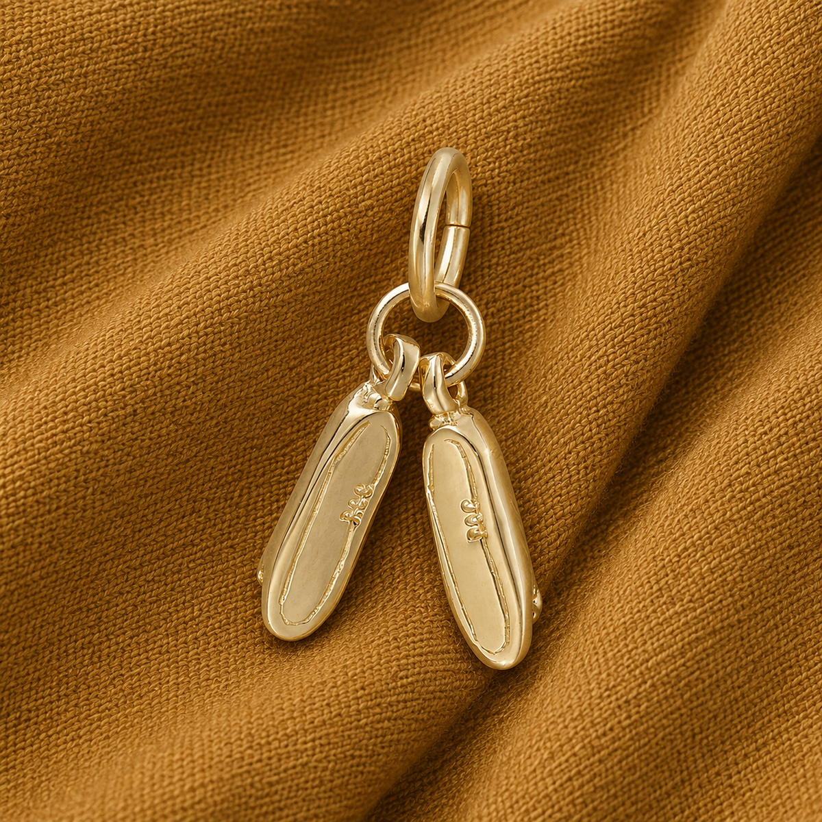 A Pair of Ballet Shoes Charm by Rembrandt Charms, gold plated and delicately engraved on the soles, rests atop a brown textured fabric background—an elegant piece reminiscent of tiny ballet shoes and a thoughtful gift for dancers.