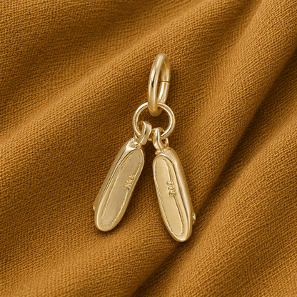 A Pair of Ballet Shoes Charm by Rembrandt Charms, gold plated and delicately engraved on the soles, rests atop a brown textured fabric background—an elegant piece reminiscent of tiny ballet shoes and a thoughtful gift for dancers.