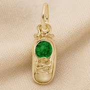 The May Baby Shoe Charm- Gold Plated by Rembrandt Charms features a round green gemstone at its center and is displayed on a beige textured fabric background—making it an ideal May birthstone charm and a thoughtful May birthday gift.