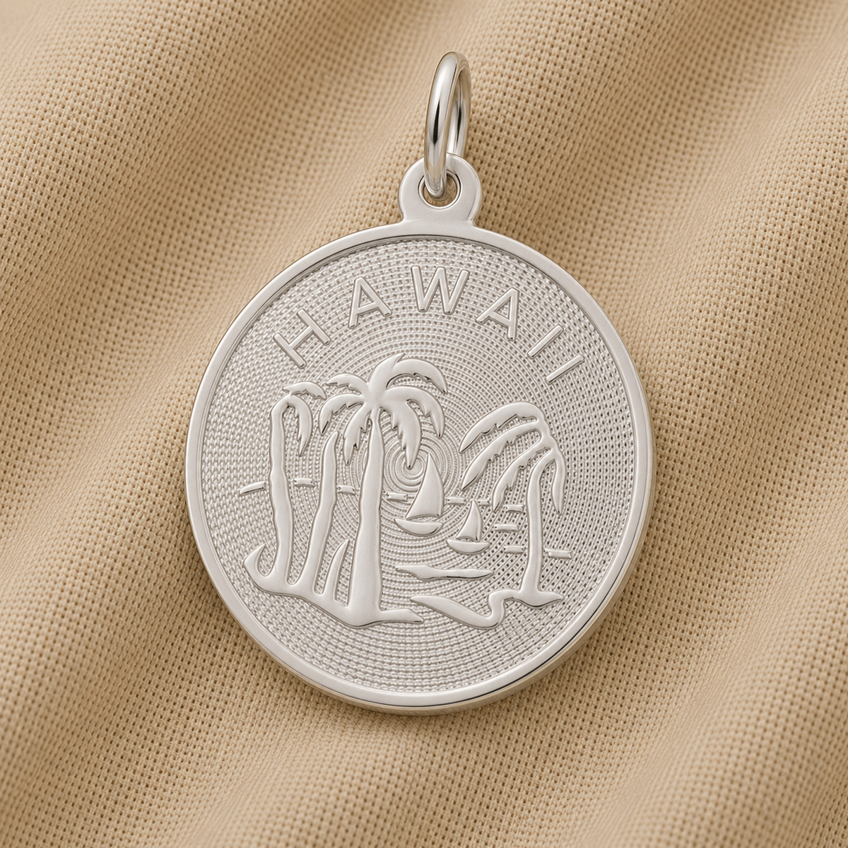 The Hawaii Palm Tree Charm in sterling silver by Rembrandt Charms, featuring “HAWAII” with engraved palm trees and a sailboat, rests on beige textured fabric—making it the perfect tropical travel keepsake and Hawaii-inspired jewelry.