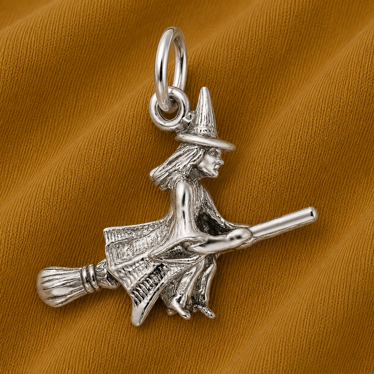 Add a touch of enchantment to your jewelry collection this spooky season with the Witch Charm - Sterling Silver by Rembrandt Charms—a beautifully crafted silver pendant featuring a witch riding a broomstick, elegantly displayed against a textured brown fabric background.