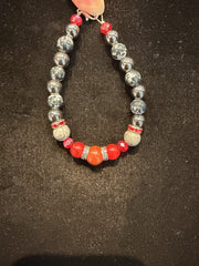 The Hemitite, Snowflake obsidion, picasso jasper, red plastic, red jasper bracelet by Redemption Jewelry features gray, silver, white, and red beads—including a large central red bead—displayed on black fabric.